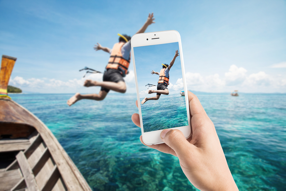 Man using photography app to take a picture of a man jumping out of a boat into the ocean